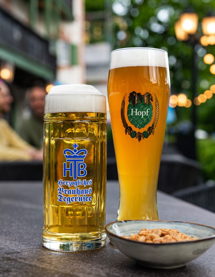 Im Biergarten des Hotel Terofal in Schliersee sieht man ein Weißbier und ein Helles. Im Hintergrund sieht man das Hotel, unscharfe Lichter und zwei Gäste die ihren Tag genießen.